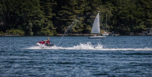 A person rides a jet ski on a lake, while a sailboat glides in the background among lush green trees.