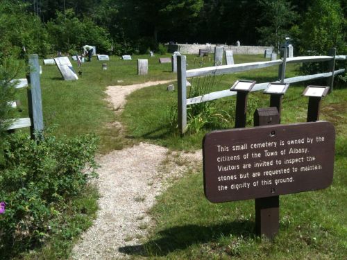 A small cemetery with a gravel path, surrounded by a wooden fence and gravestones, featuring an informational sign.