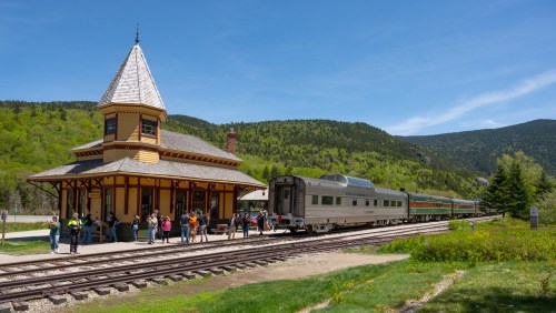 A vintage train station with a peaked roof, surrounded by greenery, as passengers board a train on a sunny day.