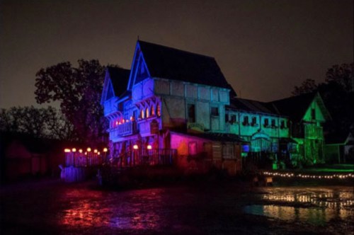 A spooky, illuminated house at night, featuring colorful lights and a reflective surface in front.