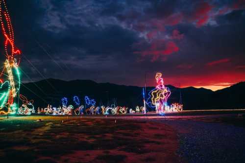 Colorful holiday lights illuminate a landscape against a dramatic sunset and darkening sky, with mountains in the background.