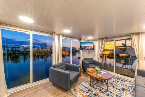 Cozy living room with two gray chairs, a round table, and large windows showcasing a marina at sunset.