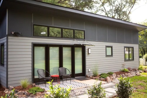 Modern gray house with large windows, a front porch, and two chairs on a patterned rug surrounded by greenery.