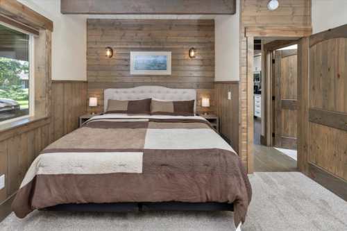 Cozy bedroom featuring a queen bed with a brown and beige quilt, wooden walls, and natural light from large windows.