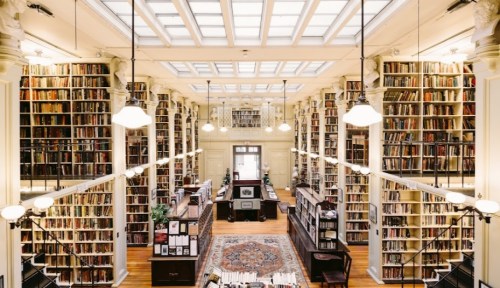 A spacious library with tall bookshelves, wooden floors, and large windows, illuminated by hanging lights.