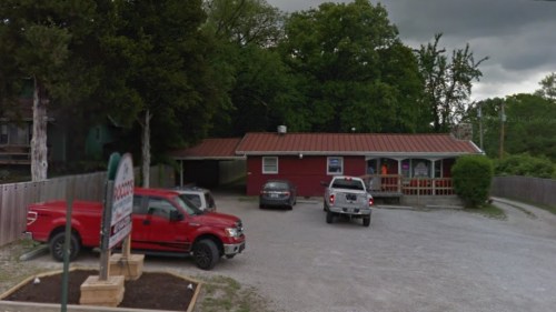 A red building with a metal roof, surrounded by trees, with parked vehicles in a gravel lot.