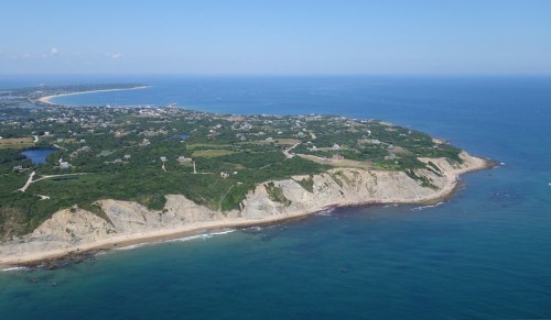 Aerial view of a coastal landscape featuring cliffs, greenery, and a clear blue ocean under a bright sky.
