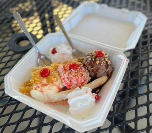A takeout container with banana split, featuring scoops of ice cream, whipped cream, cherries, and toppings.