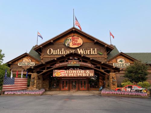 Exterior of Bass Pro Shops Outdoor World, featuring flags, a sign reading "Honor & Remember," and colorful flower beds.