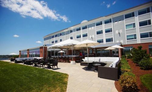 A modern hotel exterior with outdoor seating, umbrellas, and a view of the water under a clear blue sky.