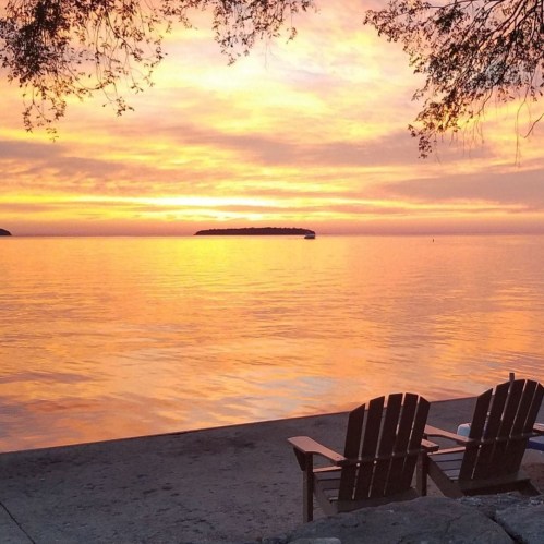 A serene sunset over water, with silhouettes of chairs on a sandy shore and an island in the distance.