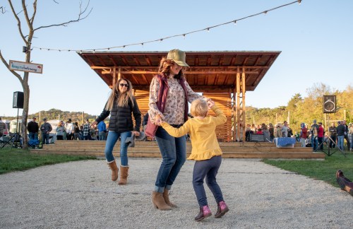 A woman dances with a young girl outdoors, while another woman watches, in a lively event setting.