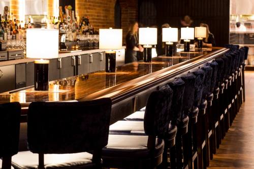 A stylish bar with a polished wooden counter, illuminated lamps, and plush seating, set in a warm, inviting atmosphere.