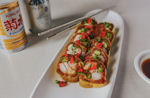 A plate of sushi rolls topped with green onions and red roe, accompanied by dipping sauce and drinks in the background.