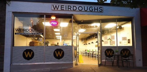Exterior of "Weirdoughs" pizzeria with an open sign, featuring a modern design and outdoor seating.
