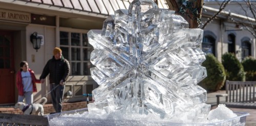 A large ice sculpture of a snowflake sits in a plaza, with people walking a dog in the background.