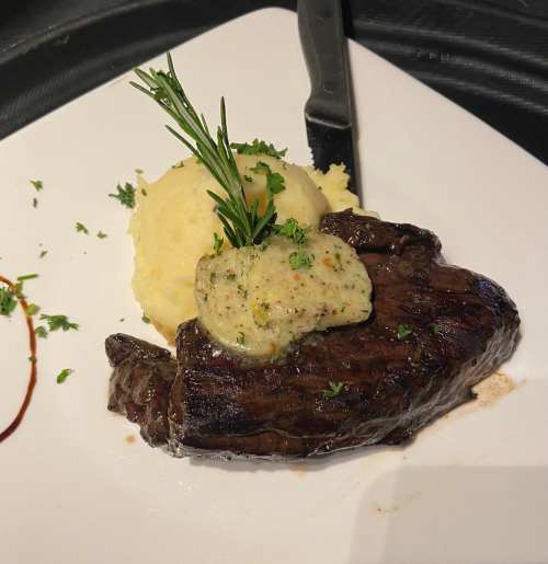 A plated steak with herb butter on top, served alongside creamy mashed potatoes and garnished with fresh herbs.