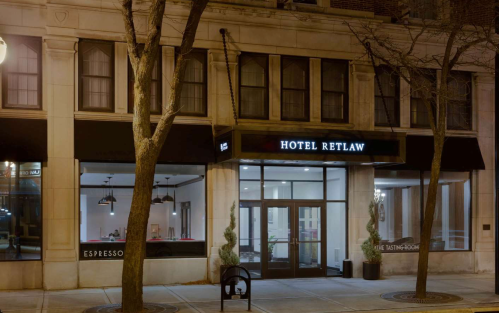Exterior view of Hotel Retlaw at night, featuring large windows and a modern entrance, surrounded by trees.
