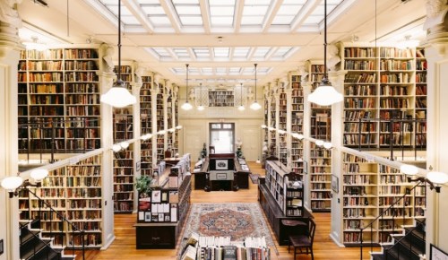 A spacious library with tall bookshelves, wooden floors, and large windows, filled with books and warm lighting.