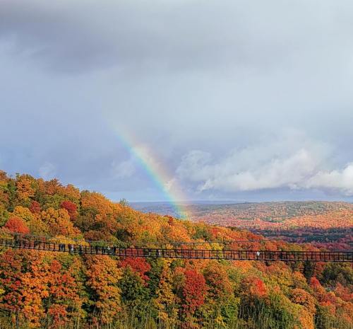 A vibrant autumn landscape with colorful trees and a rainbow arching over a valley under a partly cloudy sky.