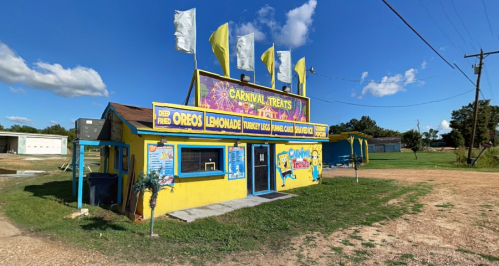 A colorful carnival treat stand with yellow flags, serving snacks like deep-fried Oreos and lemonade, set in a rural area.