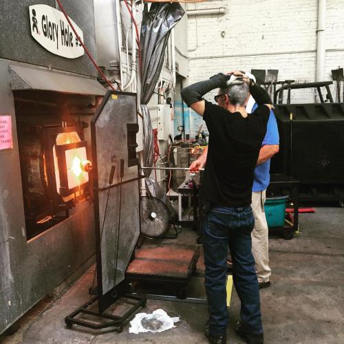 Two people working in a glassblowing studio, one shaping molten glass in a furnace.