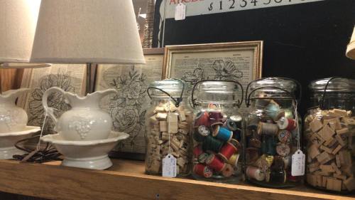 A display of jars filled with spools and wooden blocks, alongside a lamp and framed botanical prints.