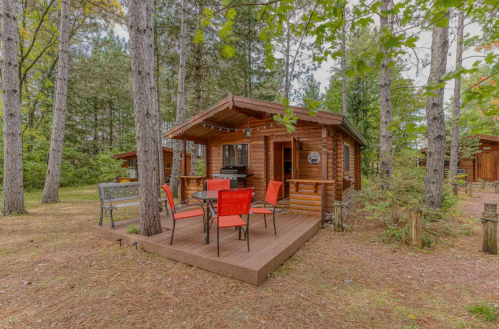 A cozy wooden cabin surrounded by tall trees, featuring a deck with red chairs and a grill.