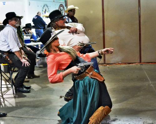 A woman in a cowboy outfit draws a gun in a shooting competition, while spectators watch in the background.