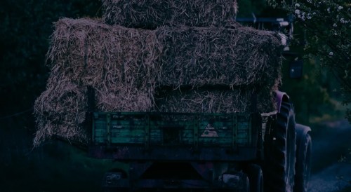 A tractor loaded with large hay bales drives down a rural path surrounded by greenery.