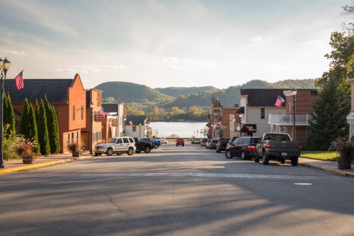 A scenic view of a small town street lined with shops, cars parked, and mountains in the background under a clear sky.