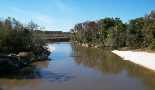 A calm river flows through a wooded area, reflecting trees and a clear blue sky. Sandy banks line the water's edge.