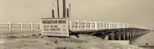 Black and white photo of a long bridge with a sign indicating tolls for cars and trucks.