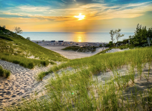 Sunset over a sandy beach, with grassy dunes in the foreground and calm waters in the background.