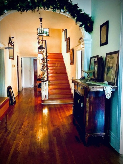 A warm, inviting hallway with wooden floors, a staircase, and vintage decor, featuring framed pictures and greenery.