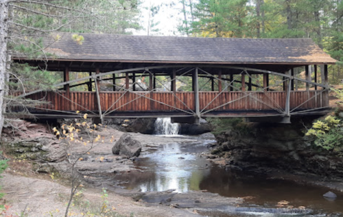 A wooden covered bridge spans a rocky stream, surrounded by trees and autumn foliage. A small waterfall is visible below.