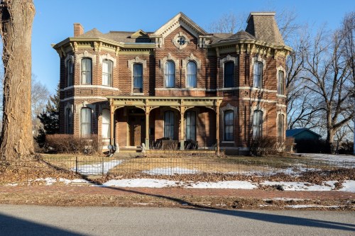 A large, historic brick house with ornate architecture, featuring a wraparound porch and snow on the ground.