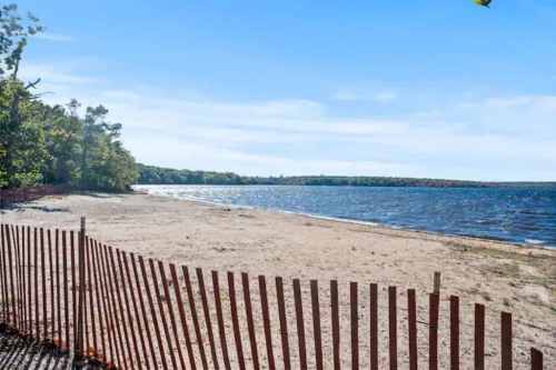 A sandy beach with a wooden fence, surrounded by trees and a calm body of water under a clear blue sky.