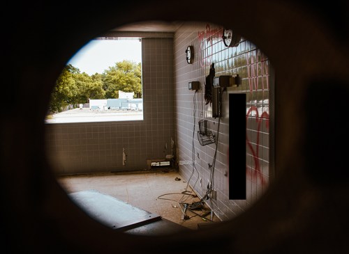 View through a circular opening into an abandoned room with graffiti, clocks, and scattered debris.