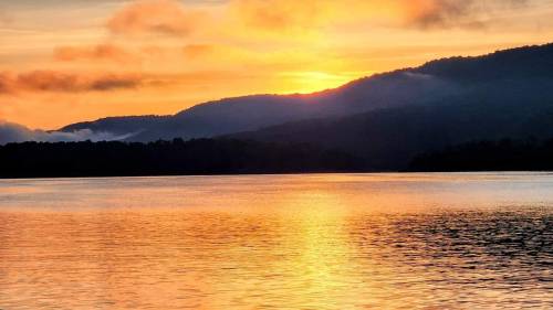 Sunset over a calm lake, with vibrant orange and yellow hues reflecting on the water and mountains in the background.