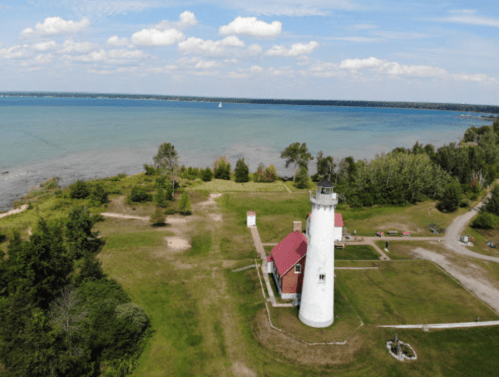 Aerial view of a white lighthouse with a red roof, surrounded by greenery and a blue lake under a cloudy sky.