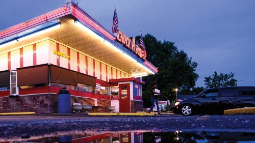 A brightly lit diner at dusk, with neon signs and a reflection in a puddle on the ground.