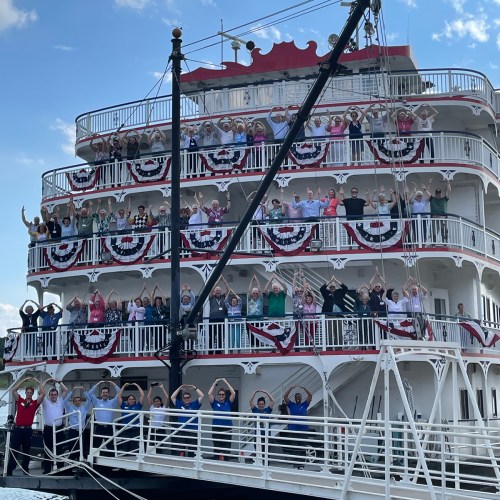 A large group of people cheerfully posing on a decorated riverboat, with multiple decks and festive banners.