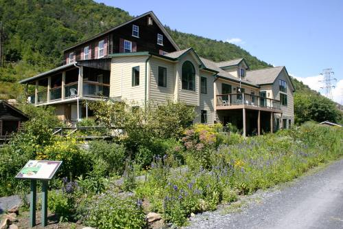 A large, multi-story house surrounded by lush greenery and colorful flowers, set against a mountainous backdrop.
