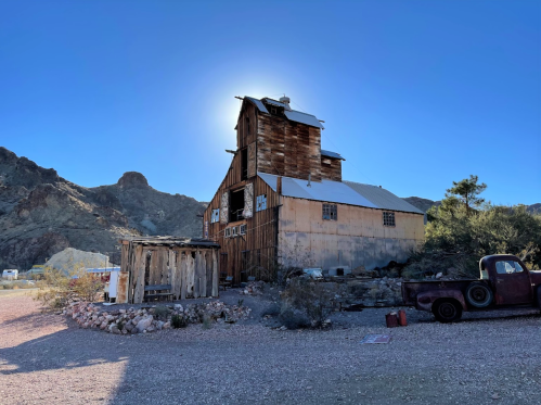 An old wooden building with a rusty roof, surrounded by mountains and a clear blue sky. A vintage truck is nearby.