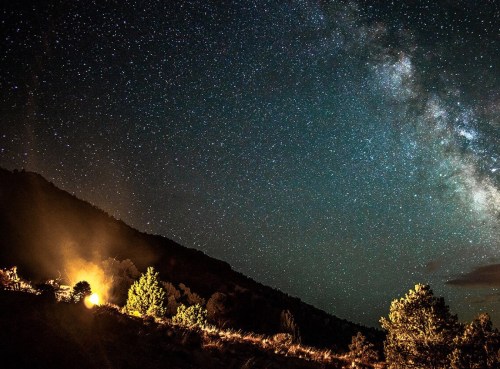 A starry night sky with the Milky Way visible above a mountain landscape, illuminated by a warm glow from below.
