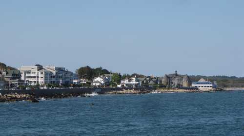 Coastal view of a shoreline with buildings, trees, and calm blue water under a clear sky.