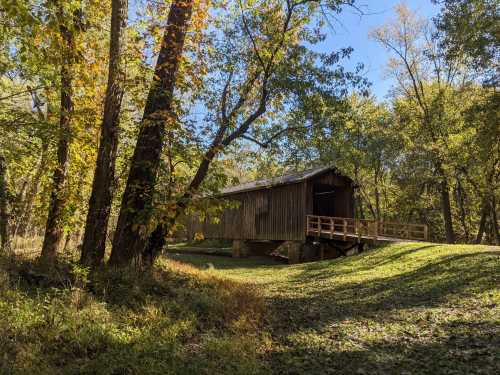 A rustic wooden covered bridge surrounded by trees with autumn foliage under a clear blue sky.