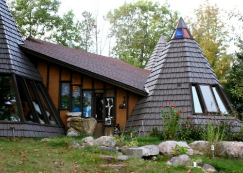 A unique house with a triangular roof, large windows, and a stone garden, surrounded by trees and greenery.