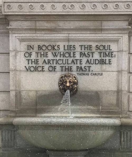A stone wall features a quote by Thomas Carlyle about books, with a lion's head fountain below it.